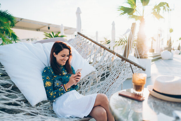 Entspannte Frau im Strandcafé, die in der Hängematte sitzt und mit Freunden Nachrichten schreibt.