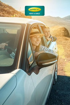 Mutter und Sohn sitzen in einem weißen Auto mit Blick auf das wunderschöne Meer. Verlinkung zu SunnyCars Angeboten mit 15 Euro Rabatt.